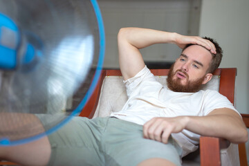 Fototapeta Young bearded man using electric fan at home, sitting on couch cooling off during hot weather, suffering from heat, high temperature obraz