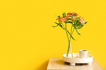 Vase with beautiful alstroemeria flowers and cup of coffee on table near yellow wall