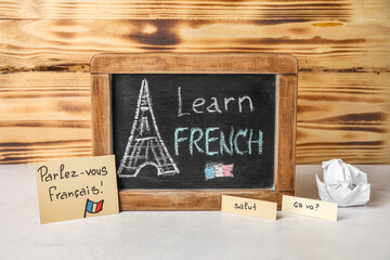 Chalkboard with text LEARN FRENCH, words and crumpled paper on table near wooden wall