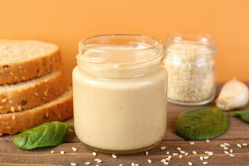Jars with tasty tahini, sesame seeds, bread and spinach on wooden table near color wall, closeup