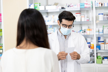 Middle eastern male pharmacist wearing protective hygienic mask to prevent infection selling medications to woman patient to prescription and making drug recommendations in modern pharmacy