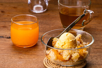 High angle view of delicious homemade peach cobbler with a scoop of vanilla ice cream and metal spoon in transparent glass bowl on bamboo mat with a glass of fresh orange juice and a cup of hot tea.