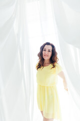 Naklejka premium beautiful brunette woman in a yellow dress stands in a bright room by the window