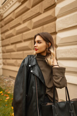 Fototapeta na wymiar Fashion young beautiful girl in fashionable casual clothes with a leather jacket and a green pullover walks on an autumn day near a vintage building