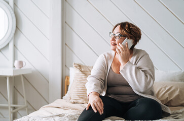 Smiling middle aged plus size woman using mobile phone on bed at home