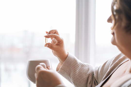 Close up photo of middle aged woman holding omega 3 capsule and mug of water in hands near window at home
