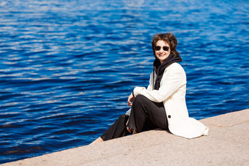 Girl sits on concrete flooring embankment in white coat and black sweatshirt in sunglasses, smiles happily on a warm sunny day. Relax in the city