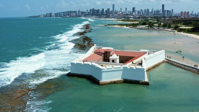 Dolly in aerial drone shot of the historic star shaped Reis Magos fort built on a reef with the coastal capital city of Natal in Rio Grande do Norte, Brazil in the background on a warm summer day