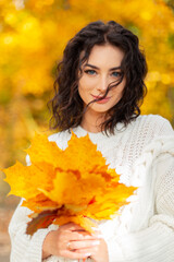 Happy beautiful young girl model with a curly hairstyle in vintage knitted sweater holds a bouquet of autumn yellow leaves and walks in a golden park