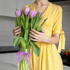 Bouquet of purple tulips in a transparent vase against the background of a woman in a white dress in the kitchen