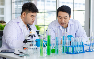Asian professional male scientist researcher in white lab coat and rubber gloves sitting using microscope inspecting quality of vegetable in laboratory while colleague typing data in tablet computer.