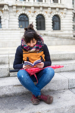 Full length of female tourist wearing warm clothing reading guidebook while sitting on steps in city