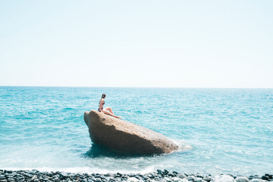 Side view of woman wearing bikini sitting on rock in sea against clear sky during sunny day