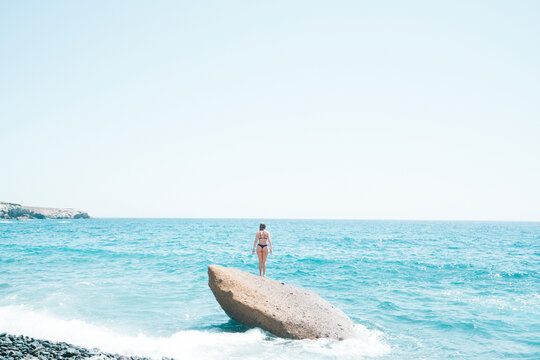 Rear view of woman wearing bikini standing on rock in sea against clear sky during sunny day