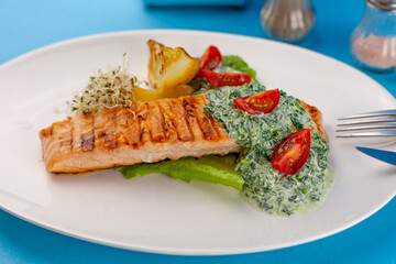 Grilled salmon steak. A piece of red fish on a white plate and a blue background on the table. Fish food with sauce, tomatoes, lemon. Restaurant menu. Grill fried