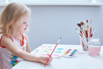A curly-haired cute girl paints at a small table in kindergarten.