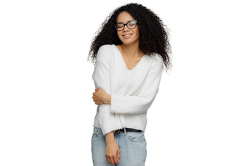 Indoor shot of sincere woman keeps hand partly crossed, dressed in white jumper and jeans, has eyes...