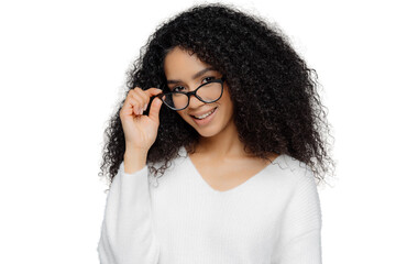 Horizontal shot of satisfied female teacher keeps hand on frame of glasses, smiles happily, has Afro hairstyle, wears casual white jumper, stands indoor over grey background. Good mood and beauty
