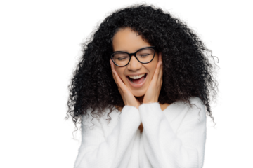 Overemotive female touches cheeks laughs from joy, keeps eyes closed, cannot stop giggling, has Afro hairstyle, dressed in white jumper, models against blue background. Ethnicity and emotions concept