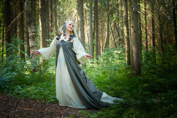Adult mature woman 40-60 in a green long fairy dress in forest. Photo shoot in style of dryad and queen of nature. Fairy who loves nature in beautiful green summer forest. Concept of caring for nature - obrazy, fototapety, plakaty