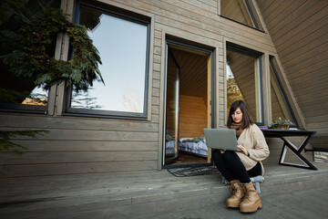 Remote work and escaping to nature concept. Woman works on laptop against tiny cabin house. - obrazy, fototapety, plakaty