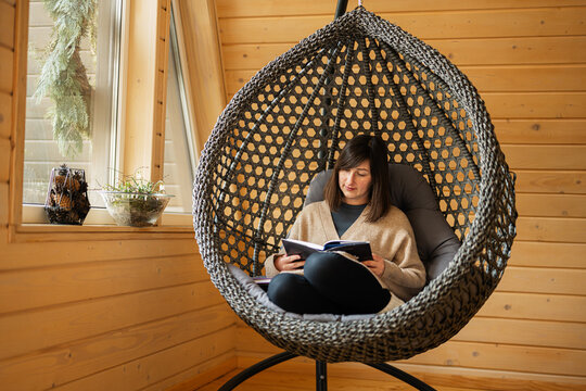 Remote work and escaping to nature concept. Woman sit in egg chair swing and read book in wooden tiny cabin house.