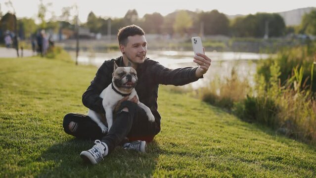 Handsome Happy Man Is Sitting on Grass Near the Local Lake with His Lovely Brooklyn Bulldog and Making Video Call to His Friends. Technology and People Dogs Friendship Concept