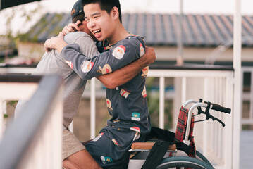  Young man with disability on wheelchair and father or volunteer or caregiver at happy time in the house, Caring for love and positive energy from parents, mental health, Family Caregiving concept.