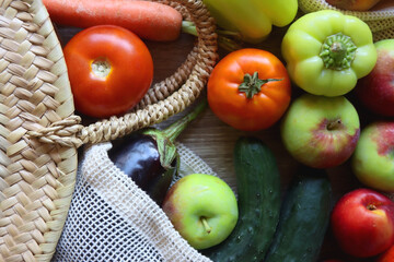 Straw bag and reusable fabric bags filled with various healthy fruit and vegetables. Wooden background, top view.
