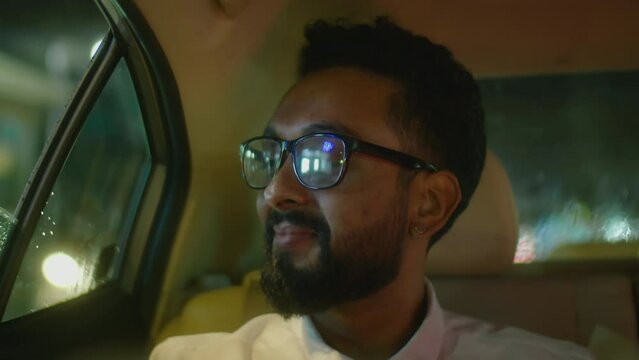 Chest up shot of cheerful businessman in formalwear and glasses looking through opened car window and smiling while riding in taxi in night city