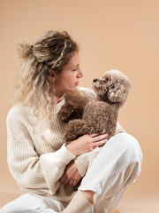Cute girl with a dog in a sweater on a beige background. Curly woman with chocolate poodle. cozy with a pet