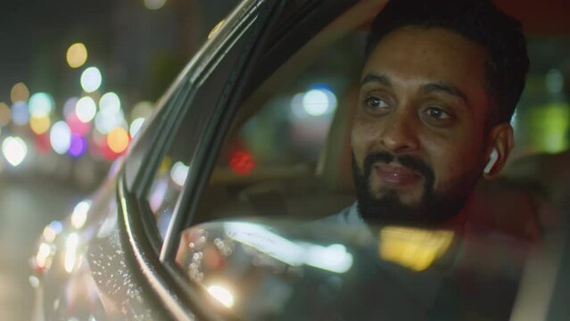 View from outside of car of businessman in wireless earphones looking through opened window and smiling while enjoying view of night city during taxi ride