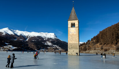 CURON VENOSTA, 06 January 2023: Tourists walk on the frozen lake of Resia. It is an artificial reservoir in the west of South Tyrol at Curon in Val Venosta, Italy