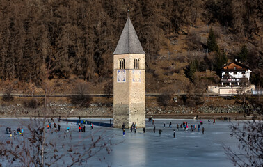 CURON VENOSTA, 06 January 2023: Tourists walk on the frozen lake of Resia. It is an artificial reservoir in the west of South Tyrol at Curon in Val Venosta, Italy