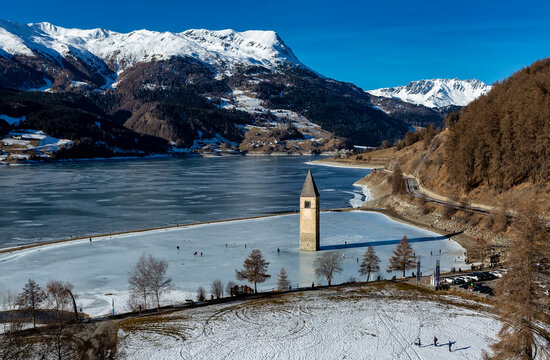 CURON VENOSTA, 06 January 2023: Tourists walk on the frozen lake of Resia. It is an artificial reservoir in the west of South Tyrol at Curon in Val Venosta, Italy