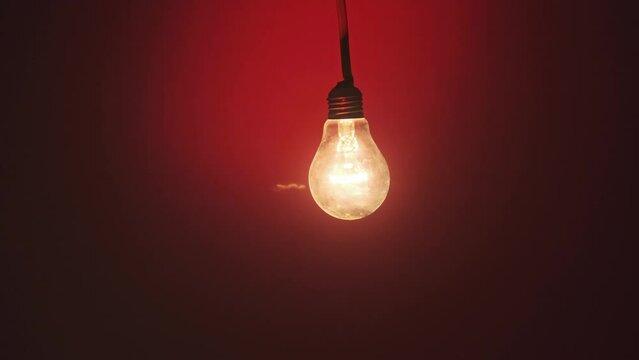 An incandescent lamp lights up when a finger touches it on dark red background. Glass bulb on wire glows, flickers, and slowly turns on and off close-up. Flashing filament. Copy space, place for text