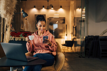 Happy African businesswoman with hair in a bun, holding a cup of coffee, and using a smartphone.