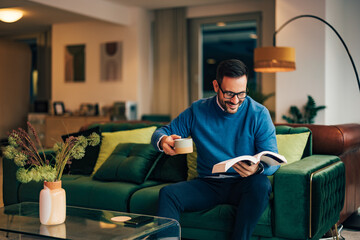 Positive businessman reading a book, holding a cup of coffee, and enjoying himself.