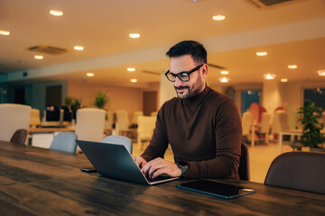 Smiling businessman typing an e-mail to clients over the laptop, sitting at the office.