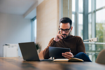 Thoughtful businessman reading something over the tablet, sitting at the workplace.