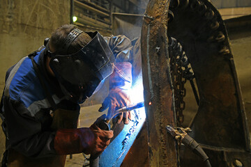 A worker welding metal parts on a construction site. A welder welds parts of a large machine in a...