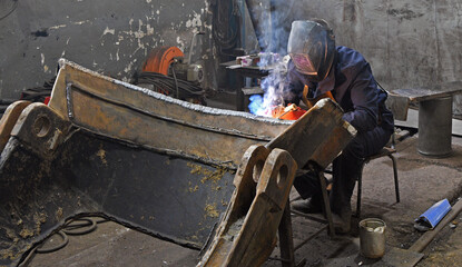 A worker welding metal parts on a construction site. A welder welds parts of a large machine in a...