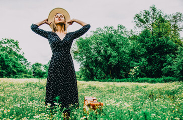 Blonde girl with suitcase and hat in chamomiles flowers field in summer time