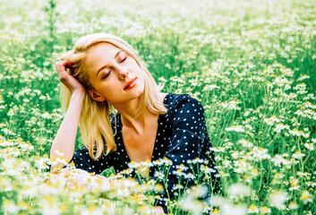 Blonde woman in black dress is sitting in countryside chamomiles flowers field