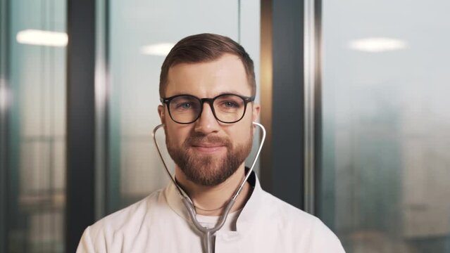 Portrait Of A Male Doctor In A White Coat Wearing A Stethoscope And Directing An Earpiece Towards The Camera Against The Backdrop Of Panoramic Windows In A Modern New Clinic