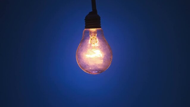 Incandescent lamp glows and flickers on dark blue background. Edison light bulb slowly turned on and off, pulsing light close-up. Warm flashing filament. Retro classic bulb. Copy space, place for text