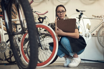 Is it the white or pink one. Full length shot of an attractive young woman crouching in her bicycle shop and using her cellphone while holding a clipboard.