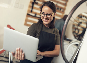 Emails are how I stay in touch with clients. Shot of an attractive young woman standing alone in her bicycle shop and using her laptop. - Powered by Adobe