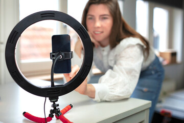 Lifestyle beauty blogger streaming online from home studio. Diverse millennial woman working as make up artist and recording video for social media on smartphone with led ring lamp, blogging content 