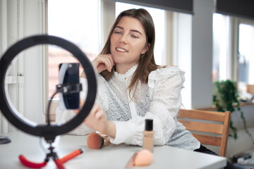 Lifestyle beauty blogger streaming online from home studio. Diverse millennial woman working as make up artist and recording video for social media on smartphone with led ring lamp, blogging content  - obrazy, fototapety, plakaty